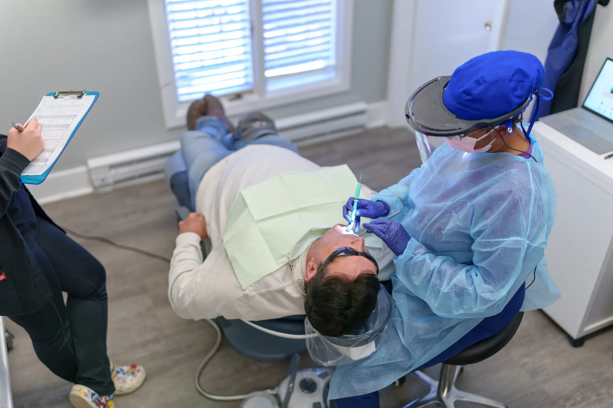White, male patient lays on a chair, receiving a check up from a multiracial, female dentist who is sitting beside him in an office.