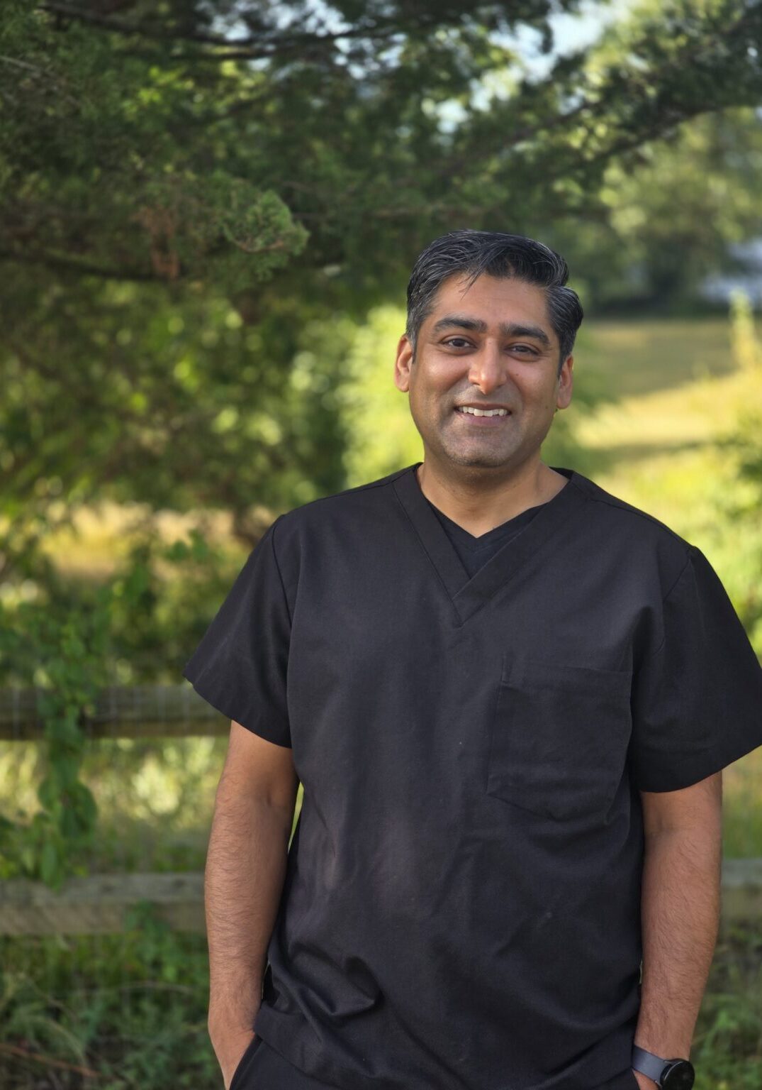 A smiling man in black scrubs stands outdoors with greenery in the background.